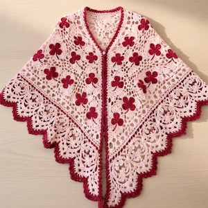 Puede incluir: Un chal de crochet hecho a mano, con frente abierto, en un tono rosa claro, adornado con motivos de tréboles rojos oscuros. El chal presenta un borde festoneado y un borde decorativo, creando un diseño delicado y femenino. Es un accesorio acogedor.
