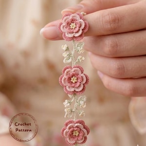 Puede incluir: Primer plano de un diseño floral de ganchillo. El diseño presenta tres flores rosas y crema con centros amarillos, conectadas por un tallo con pequeñas hojas verdes. El texto "Crochet Pattern" está en un marco circular.