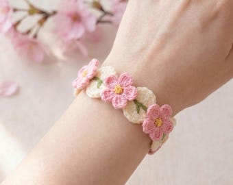Puede incluir: Una pulsera de ganchillo hecha a mano con diseños de flores rosas y hojas color crema. La pulsera está adornada con flores rosas con centros amarillos y tallos verdes, creando un accesorio delicado y femenino.