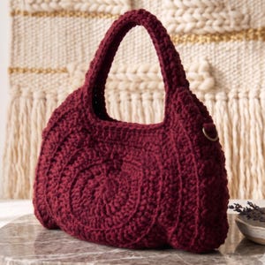 Peut inclure: Un sac à main bordeaux profond réalisé en fil épais et texturé selon une technique de crochet. Le sac présente un motif circulaire arrondi sur le panneau avant, une anse intégrée unique et un petit anneau en D métallique sur le côté.