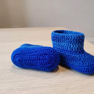 Peut inclure: Une paire de chaussons au crochet bleu vif. Un chausson est positionné debout, montrant un design montant avec un motif de points texturé, tandis que l'autre chausson est posé sur le côté, révélant la semelle en crochet solide, arrondie et côtelée.