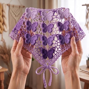 Peut inclure: Un châle en crochet violet fait à la main avec un motif de papillon. Le châle est tenu par des mains, mettant en valeur le travail ajouré complexe et les délicates applications de papillons. La palette de couleurs comprend diverses nuances de violet.