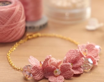 Puede incluir: Un delicado collar de flores de ganchillo rosa con una cadena dorada. El collar presenta varias flores de ganchillo y cuentas de cristal transparente. El fondo incluye hilo rosa y otros suministros de artesanía, lo que sugiere un artículo hecho a mano.