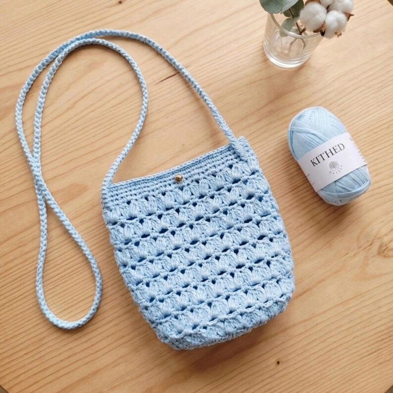 Puede incluir: Un bolso bandolera de ganchillo azul claro con una correa larga. El bolso tiene un cierre de bot&oacute;n y un dise&ntilde;o de tejido abierto. Una madeja de hilo azul claro con una etiqueta est&aacute; al lado del bolso. El bolso est&aacute; sobre una superficie de madera.