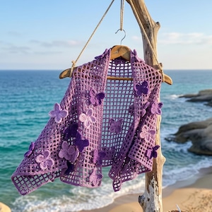 Peut inclure: Un châle en crochet lavande avec un motif quadrillé, orné de fleurs et de papillons en crochet violets de différentes nuances. Le châle est présenté sur un cintre en bois, suspendu par une corde, avec la mer et la plage en arrière-plan.