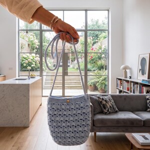 Puede incluir: Bolso de ganchillo azul claro con una correa larga y un bot&oacute;n de madera. El bolso tiene un patr&oacute;n de tejido abierto y texturizado. El bolso se sostiene frente a una ventana con vistas a un jard&iacute;n.