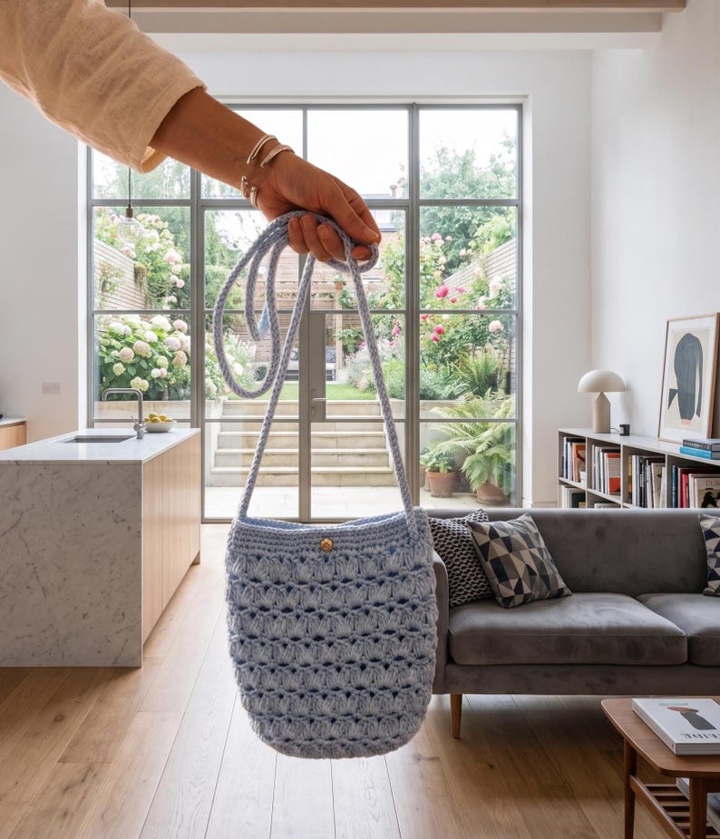 Puede incluir: Bolso de ganchillo azul claro con una correa larga y un bot&oacute;n de madera. El bolso tiene un patr&oacute;n de tejido abierto y texturizado. El bolso se sostiene frente a una ventana con vistas a un jard&iacute;n.