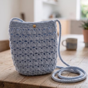 Puede incluir: Un bolso bandolera de ganchillo azul claro con una correa larga. El bolso tiene un patrón texturizado y un pequeño cierre de botón redondo de color dorado. El bolso está sobre una mesa de madera.
