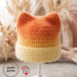 Peut inclure: Un bonnet au crochet duveteux de style ombré avec deux oreilles pointues en forme de chat sur le dessus. Le bonnet passe d'un orange profond au sommet à un jaune doré au milieu, finissant par un bord crème pâle. Inclut une icône de modèle PDF.
