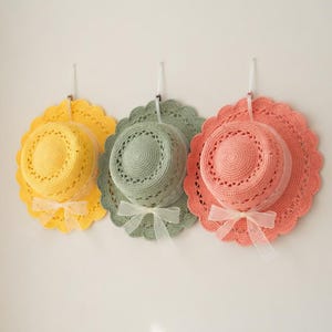 Op de afbeelding: Drie gehaakte zonnehoeden in geel, saliegroen en koraalroze hangen aan een witte muur. Elke hoed heeft een geschulpte rand, een bijpassend lint en een doorzichtig plastic schild. De hoeden zijn een charmant, decoratief item.
