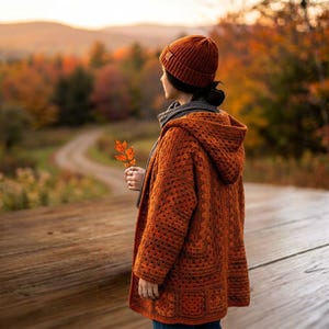 Peut inclure: Un gilet à capuche en crochet de couleur rouille avec une texture en maille à motifs carrés, associé à un bonnet en maille côtelée assorti. Le vêtement est porté sur une écharpe gris foncé, et une petite feuille orange est tenue à la main.