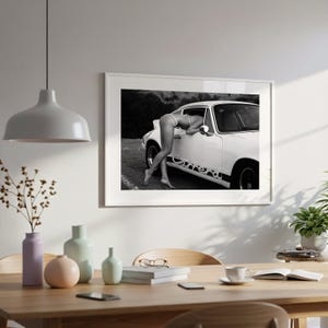 a framed black and white photograph of a person leaning out of a car window, with a dining table and chairs in the foreground.