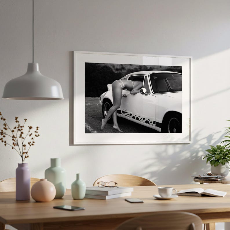a framed black and white photograph of a person leaning out of a car window, with a dining table and chairs in the foreground.