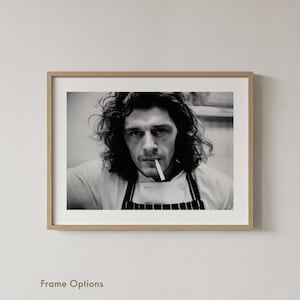 May include: Monochrome framed print featuring a person in a chef's uniform, smoking a cigarette. The person has long, curly hair and is looking directly at the viewer. The frame is a light wood colour.