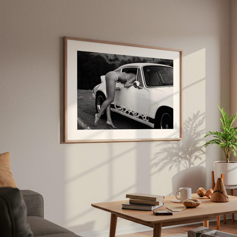 a framed black and white photograph of a woman leaning out of a car window, with a potted plant and other objects in the foreground.
