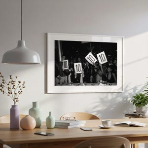 a framed black and white photograph of a group of people holding up signs with the numbers "10" and "10" on them, suggesting a competition or event. The photograph is displayed on a wall above a wooden dining table, which has various decorative items such as vases, a potted plant, and a book.