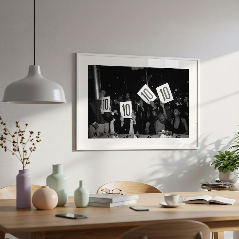 a framed black and white photograph of a group of people holding up signs with the numbers "10" and "10" on them, suggesting a competition or event. The photograph is displayed on a wall above a wooden dining table, which has various decorative items such as vases, a potted plant, and a book.