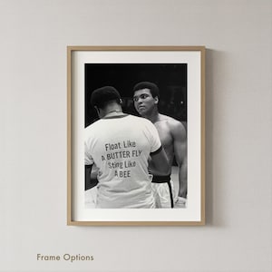 以下が含まれることがあります： 2人の男性が写った、額縁に入った白黒写真。一人は「Float Like A BUTTER FLY Sting Like A BEE」と書かれた白いTシャツを着ています。もう一人はカメラを見ています。写真は薄茶色のフレームに入っています。