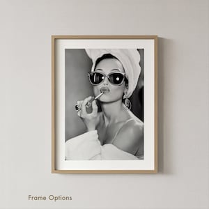 Op de afbeelding: Zwart-wit ingelijste print van een vrouw die lippenstift aanbrengt. Ze draagt een zonnebril, een handdoek op haar hoofd en een badjas. De print zit in een lichtgekleurde houten lijst. De tekst "Frame Options" staat onderaan.
