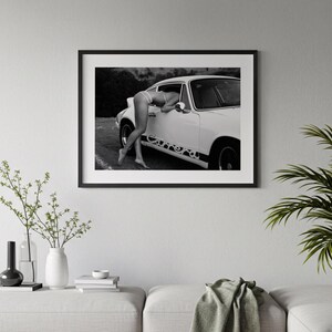 A framed black and white photograph of a person leaning out of a car window, with a potted plant and books on a table below the frame.