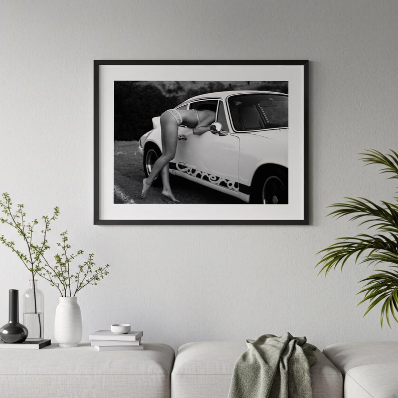 A framed black and white photograph of a person leaning out of a car window, with a potted plant and books on a table below the frame.