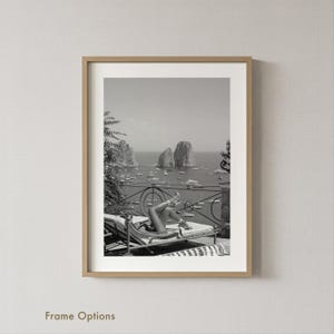 May include: Black and white framed print of a person reclining on a lounge chair overlooking a seascape. The image features a scenic view of the ocean with boats and rock formations. The frame is a light brown color.