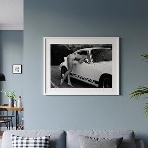 A framed black and white photograph of a person leaning out of a car window, with a couch and other decor visible in the background.