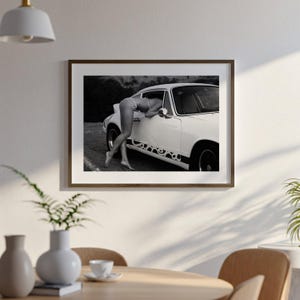 A framed black and white photograph of a person leaning out of a car window, with a potted plant and other decorative items visible in the foreground.