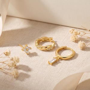 May include: A collection of gold-colored earrings. The set includes a stud earring with a four-petal design, a twisted hoop earring, and a hoop earring with a rectangular charm. All earrings are adorned with small, clear gemstones.