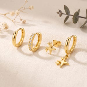May include: A set of four gold-toned earrings consisting of one plain hoop, one hoop encrusted with clear stones, one star-shaped stud earring, and one hoop earring featuring a dangling four-pointed star charm with a polished metallic finish.