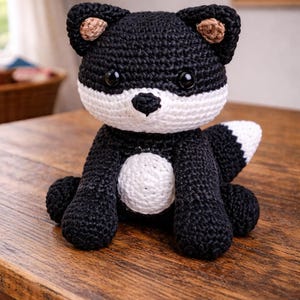 May include: A handmade crochet fox toy in black and white, with a white chest and tail tip. The fox has small, dark eyes and brown accents on its ears. It sits on a wooden surface.