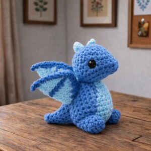 May include: A handmade blue crochet dragon with light blue wings and belly, sitting on a wooden table. The dragon has large black eyes and small horns. The wings have a striped pattern. The background includes a window, artwork, and a basket of yarn.
