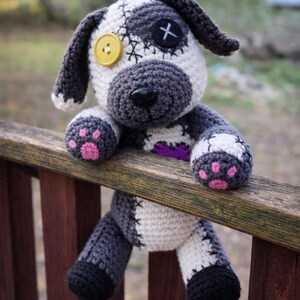 May include: Crocheted dog plushie with a patchwork design. The dog has a grey snout, ears, and paws with pink paw pads. It has a yellow button eye and a black cross stitched on the other eye. The dog is sitting on a wooden railing.