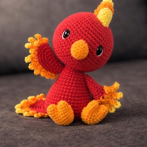 May include: A handmade crochet bird in red, orange, and yellow. The bird has a round head, a small beak, and large black eyes. Its wings and tail are detailed with fringed edges. The bird is sitting on a gray surface.