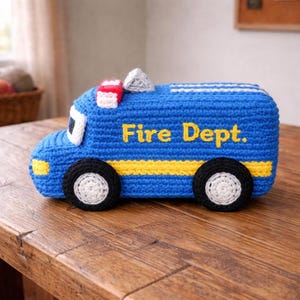 May include: A blue crocheted toy fire truck with the text "Fire Dept." in yellow. The truck has black wheels with white centers, yellow stripes, and a red and white light on top. The toy is sitting on a wooden surface.