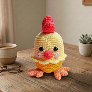 Può includere: Figurina di pollo all'uncinetto fatta a mano. Il corpo giallo ha una cresta e un becco rossi, con occhi neri e guance rosa. Il pollo ha zampe arancioni e una base gialla a balze. La statuetta è appoggiata su una superficie di legno.