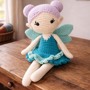 May include: A handmade crochet fairy doll with a cream-colored face and limbs, lavender hair buns, and turquoise wings. The doll wears a blue dress with a ruffled hem and is seated on a wooden surface.