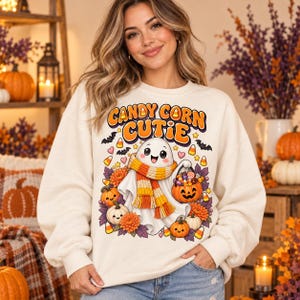 Puede incluir: Una sudadera de cuello redondo color crema con un estampado gr&aacute;fico de un fantasma sonriente que lleva una bufanda a rayas naranja, amarilla y blanca. El texto dice CANDY CORN CUTIE, rodeado de ilustraciones de calabazas y dulces.