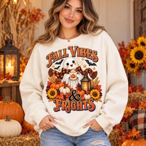Puede incluir: Una sudadera de cuello redondo color crema con un estampado gr&aacute;fico de un fantasma con bufanda de cuadros que sostiene una taza de calabaza. El dise&ntilde;o incluye el texto "FALL VIBES & FRIGHTS" rodeado de calabazas, girasoles, hojas de oto&ntilde;o y un farol.