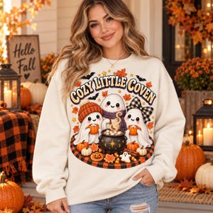 Puede incluir: Una sudadera de cuello redondo color crema con un estampado gr&aacute;fico de tres fantasmas alrededor de un caldero con el texto "COZY LITTLE COVEN". El dise&ntilde;o incluye calabazas naranjas, hojas de oto&ntilde;o, un lazo a cuadros y un gorro de punto.