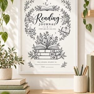 May include: A white framed print featuring black line art of a reading journal cover. The design includes stacks of books, a teacup, flowers, and the text Reading JOURNAL A little space for every story, THIS JOURNAL BELONGS TO, and YEAR.