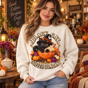 Puede incluir: Una sudadera de cuello redondo color crema con un gr&aacute;fico de un gato negro con sombrero de bruja dentro de una taza de caf&eacute; naranja. El dise&ntilde;o incluye calabazas, hojas de oto&ntilde;o y el texto Black Cat Coffee Company en letra decorativa.