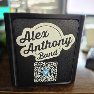 Könnte beinhalten: Ein schwarzer, quadratischer Aufsteller mit einem weißen, erhabenen Logo, das in Schreibschrift Alex Anthony Band liest. Unter dem Text befindet sich ein quadratischer, schwarz-weißer QR-Code mit einem kleinen blauen Kreissymbol und einem weißen v.
