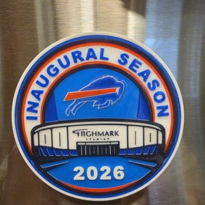 Può includere: Un magnete rotondo con un design blu, bianco e arancione. Il magnete presenta il testo "INAUGURAL SEASON" e "HIGHMARK STADIUM 2026". Il centro presenta un logo di bufalo blu.
