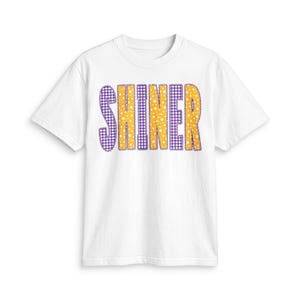 以下が含まれることがあります： 胸元にSHINERの文字がプリントされた白い半袖Tシャツ。文字は紫と白のギンガムチェック柄と、小さな白い星が散りばめられた黄色い背景が交互に配置されており、全体が紫色のスカラップ状の縁取りで囲まれています。