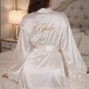 May include: Ivory satin robe with lace trim and a matching belt. The word "Bride" is embroidered in gold script on the back. The robe has long sleeves and a floral lace trim along the hem and sleeve cuffs.