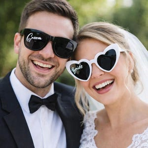 May include: A smiling couple wearing sunglasses. The groom's sunglasses are black with "Groom" in white script. The bride's sunglasses are heart-shaped and white, with "Bride" in white script.