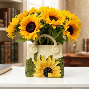 May include: A cream-colored ceramic vase shaped like a handbag, featuring a raised, sculpted sunflower design on the front with green leaves. The vase is filled with a large bouquet of bright yellow sunflowers with dark brown centers.