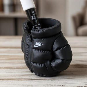 May include: A black puffer-style pen holder shaped like a miniature jacket with a white Nike logo on the front. A black and white marker with a 0.9-1.0mm nib is inserted into the top opening of the holder, which features a zipper detail.