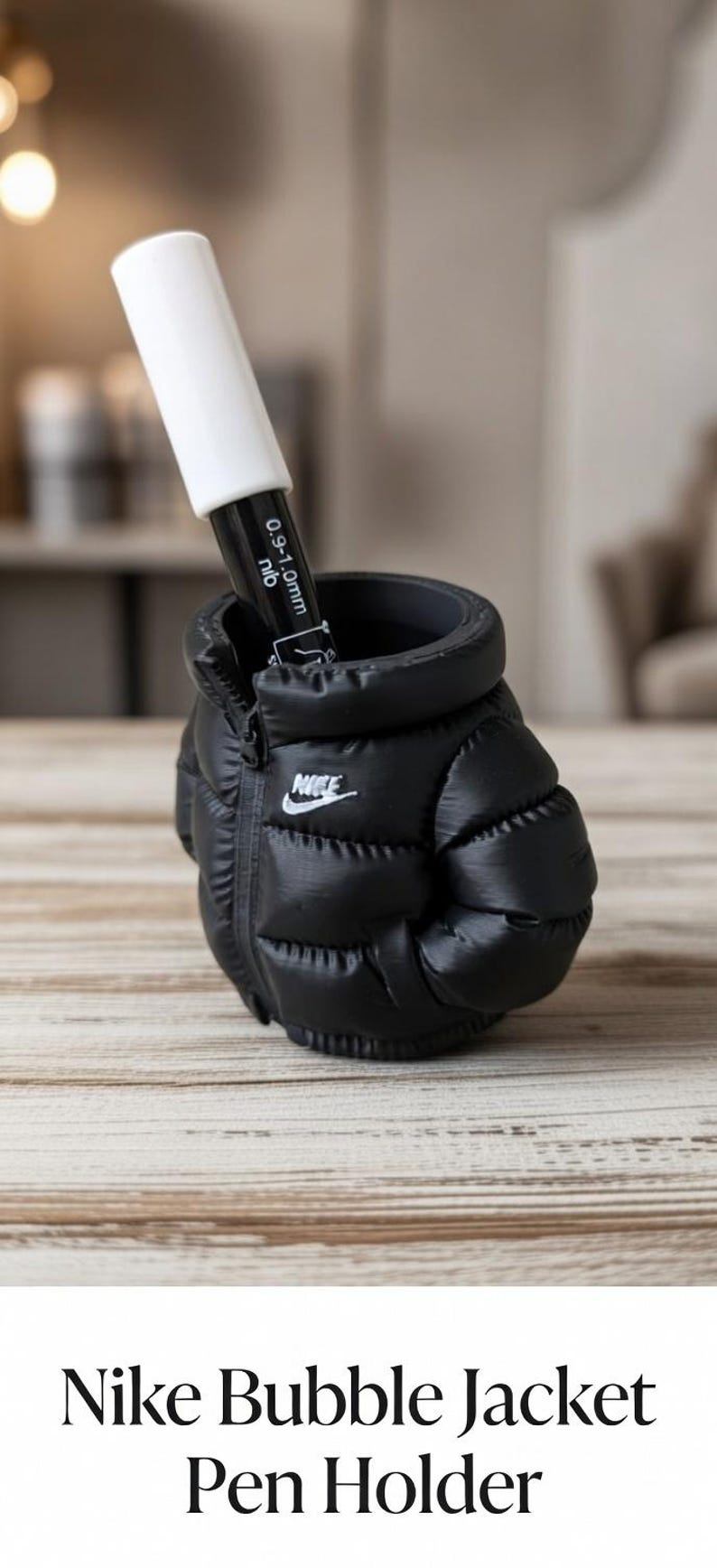 May include: A black puffer-style pen holder shaped like a miniature jacket with a white Nike logo on the front. A black and white marker with a 0.9-1.0mm nib is inserted into the top opening of the holder, which features a zipper detail.